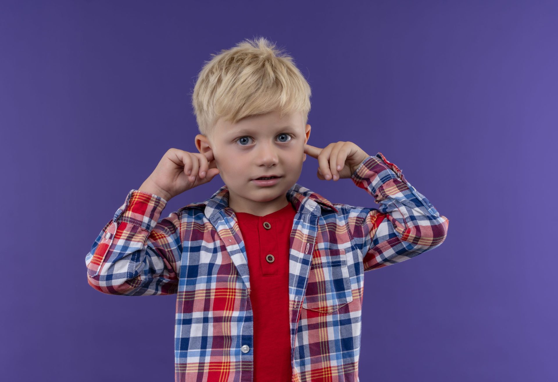 a cute little boy with blonde hair wearing checked shirt keeping hand on ears on a purple background